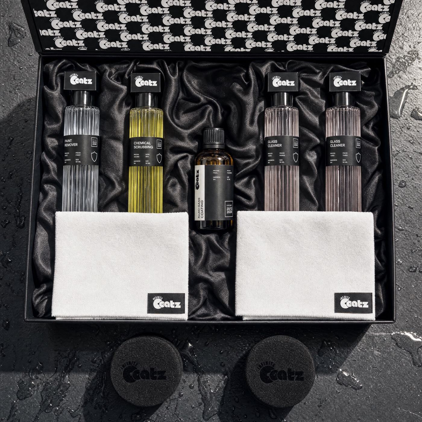 The full kit laid out on wet dark slate: five bottles, two microfiber cloths and two foam application pads beside the open box.